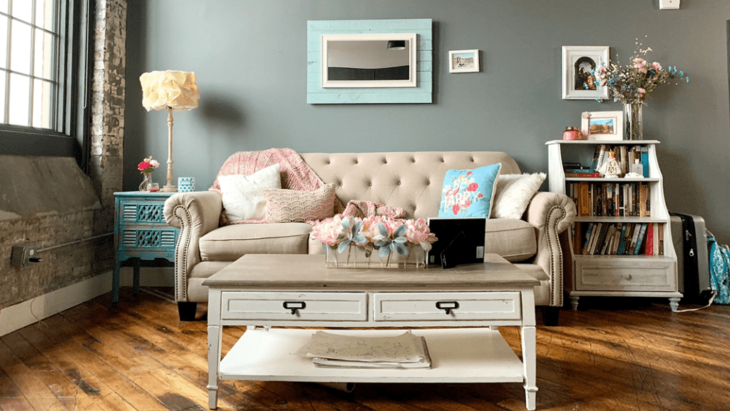 A livingroom with an off white sofa and pink and blue spring themed pillows, an off white coffee table with a pink flower arrangement