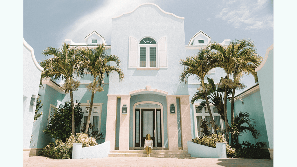 Large light blue house with palm trees and a young woman seated on the front stairs